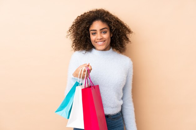Young African American woman on beige wall holding shopping bags and thinking