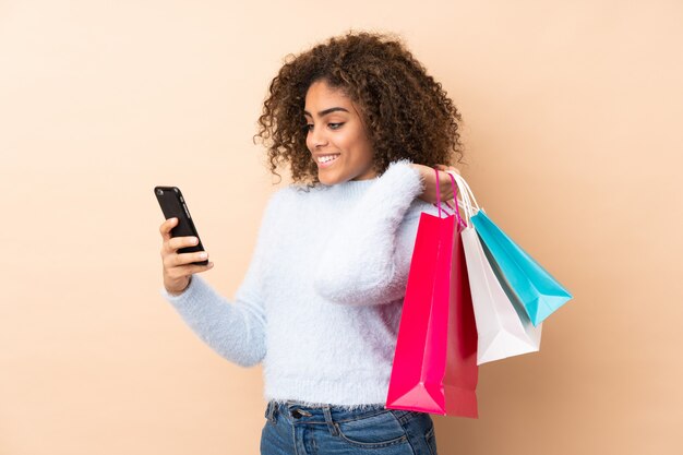 Young African American woman on beige wall holding shopping bags and writing a message with her cell phone to a friend