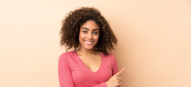 Young African American woman on beige wall pointing to the side