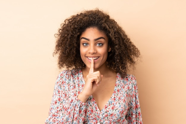 Young African American woman on beige wall showing a sign of silence gesture putting finger in mouth