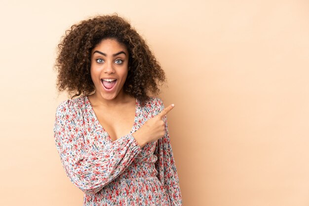 Young African American woman on beige wall surprised and pointing side