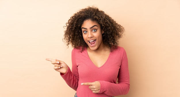 Young African American woman on beige wall surprised and pointing side