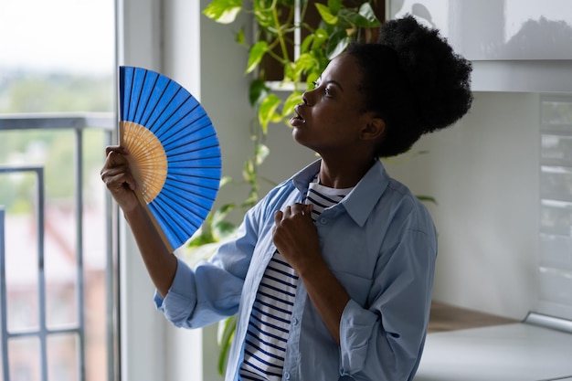 더운 날씨 때문에 손으로 부채를 부채질하는 파란색 셔츠를 입은 젊은 아프리카계 미국인 여성