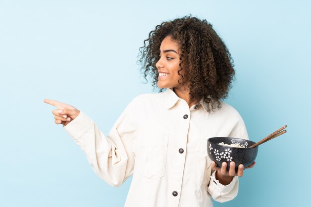 Young African American woman on blue wall pointing to the side while holding a bowl of noodles with chopsticks