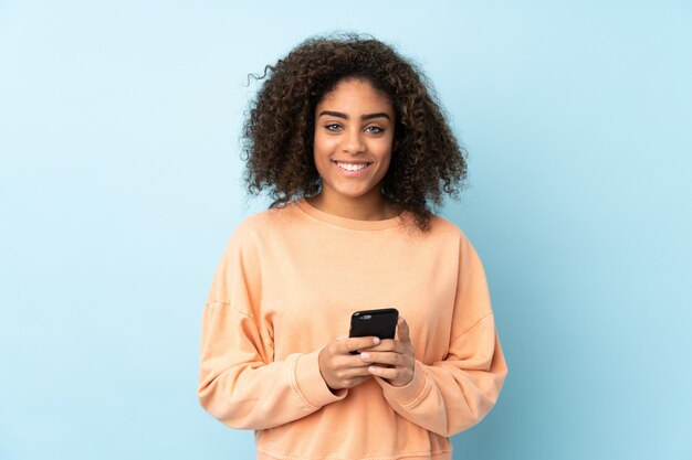 Young African American woman on blue wall sending a message with the mobile