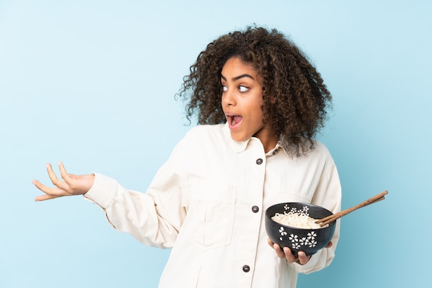 Giovane donna afroamericana sulla parete blu con espressione facciale sorpresa mentre si tiene una ciotola di noodles con le bacchette