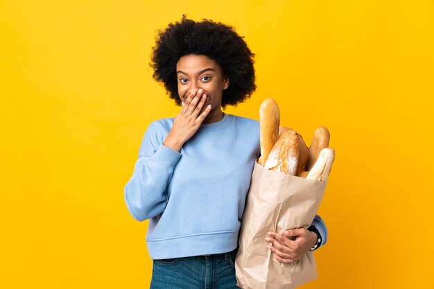 Молодая афро-американская женщина, покупающая что-то хлеб, изолирована на желтом счастливом и улыбающемся, закрывая рот руками