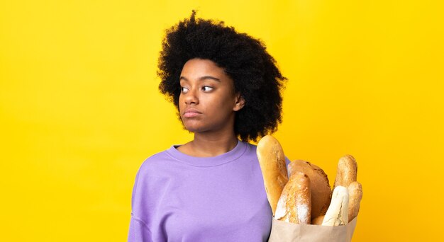 Young African American woman buying something bread isolated on yellow looking to the side