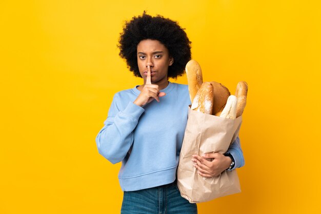 Giovane donna afro-americana che compra qualcosa di pane isolato su giallo che mostra un segno di silenzio gesto mettendo il dito in bocca