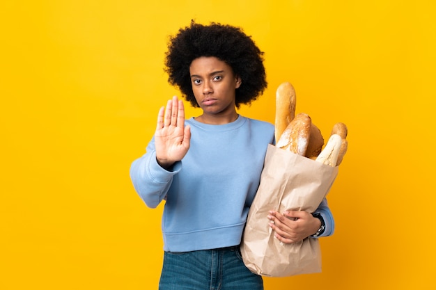 Giovane donna afroamericana che compra qualcosa di pane isolato sulla parete gialla che fa gesto di arresto