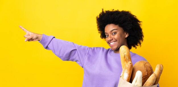 Young african american woman buying something bread isolated on yellow wall pointing finger to the side and presenting a product