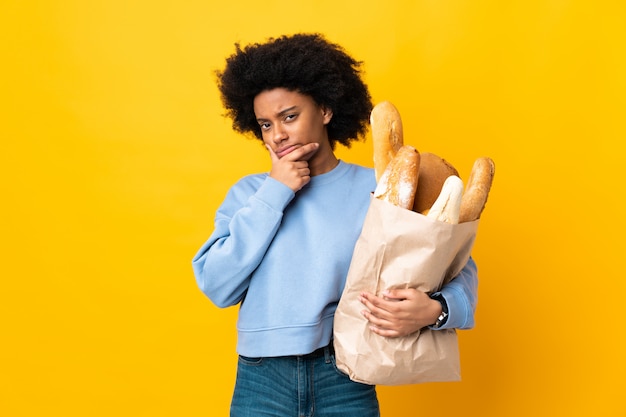 Young African American woman buying something bread on yellow wall thinking