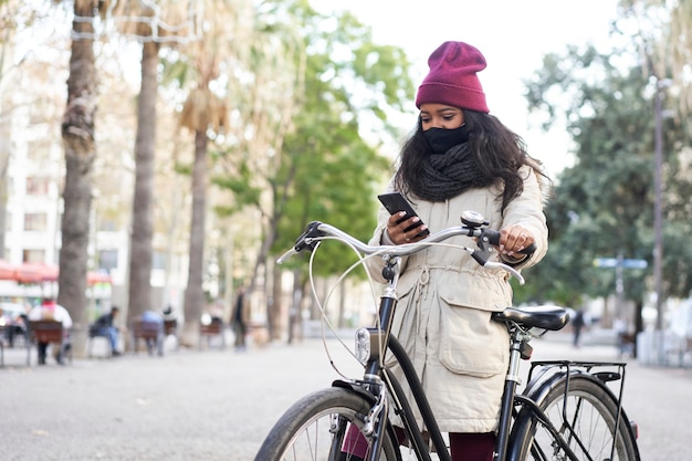 彼女の自転車でスマートフォンを使用して都市の若いアフリカ系アメリカ人の女性。彼女はスカーフとウールの帽子をかぶった冬服を着ています。