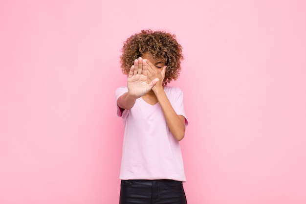 Young african american woman covering face with hand and putting other hand up front to stop camera, refusing photos or pictures against pink wall