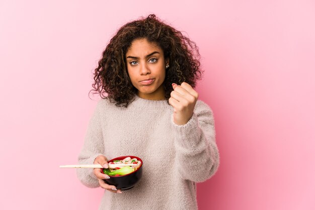 Young african american woman eating noodles showing fist to camera, aggressive facial expression.