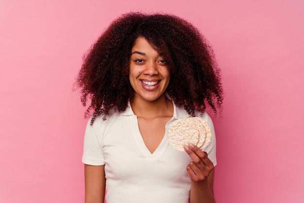 ピンクの背景に孤立した餅を食べる若いアフリカ系アメリカ人女性は、幸せで笑顔で陽気です。