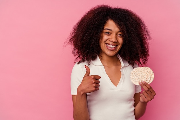 Young african american woman eating a rice cakes isolated on pink background smiling and raising thumb up