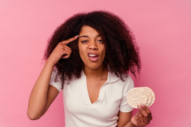 Молодая афро-американская женщина ест рисовые лепешки, изолированные на розовой стене, показывая жест разочарования указательным пальцем.