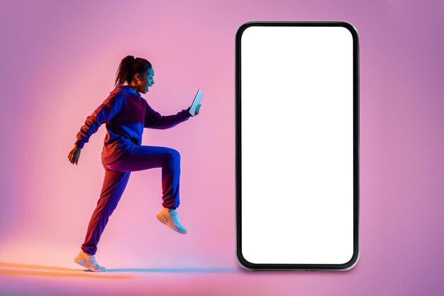 Young african american woman exercising running with cellphone next to giant smartphone with blank screen mockup