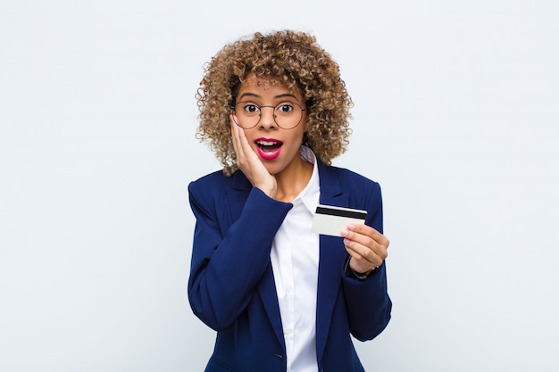 Young african american woman feeling shocked and scared, looking terrified with open mouth and hands on cheeks with a credit card