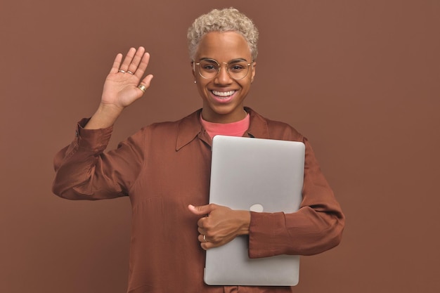 Young african american woman freelancer waving hand in greeting holding laptop