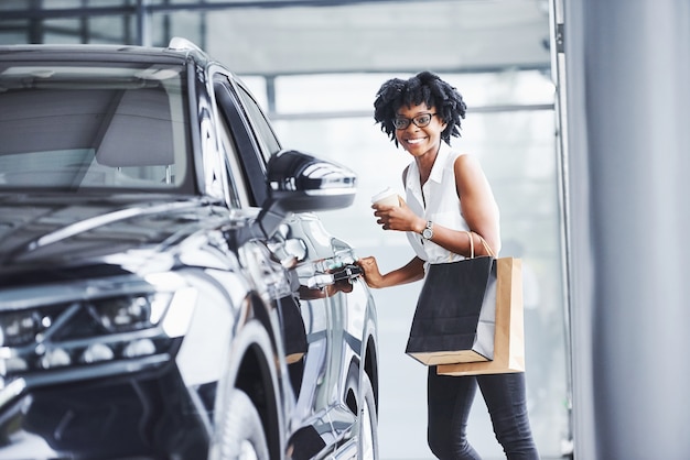 Young african american woman in glasses stands in car salon near vehicle with package in hands.