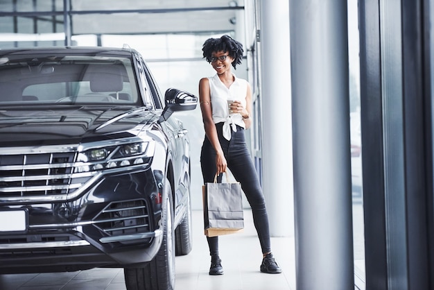 Young african american woman in glasses stands in car salon near vehicle with package in hands.