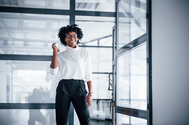 Young african american woman in glasses stands indoors in the office with phone.