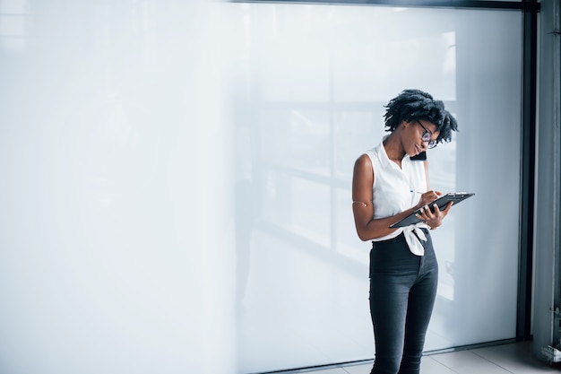 Young african american woman in glasses stands indoors in the office with tablet in hands.