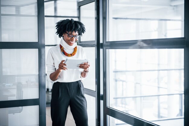 Young african american woman in glasses stands indoors in the office with tablet in hands.