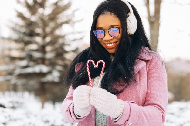 Молодая африканская американка в очках и теплых варежках держит две сладкие конфеты в форме сердца