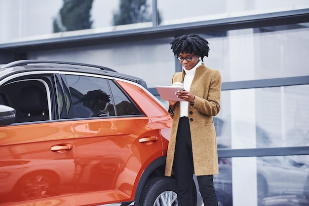 Young african american woman in glasses and with cup of drink stands outdoors near modern car.