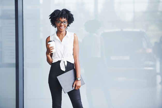 Photo young african american woman in glasses with notepad and cup of drink in hands stands indoors.