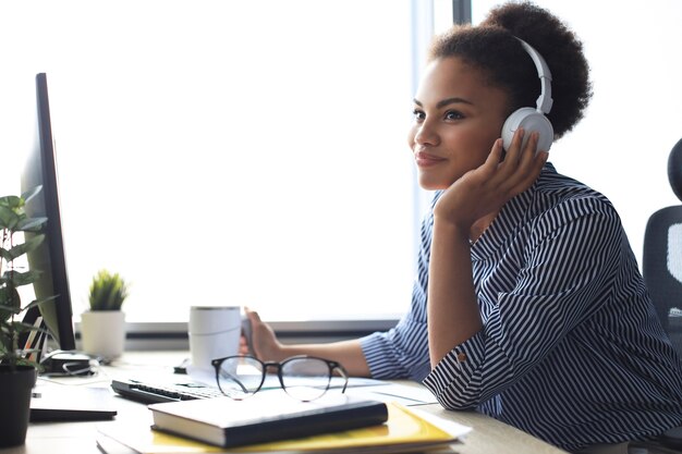 Young african american woman having a break and listening music in headphones sitting on working place.