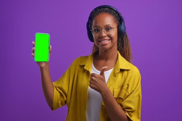 Young african american woman in headphones holding phone and showing thumbs up