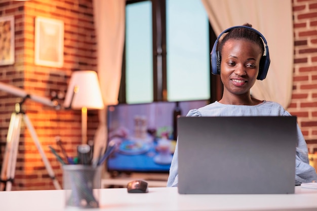 Молодая афроамериканка в наушниках дома смотрит видео на ноутбуке. Улыбающаяся студентка посещает дистанционный урок в университете, учится с компьютером в современной комнате