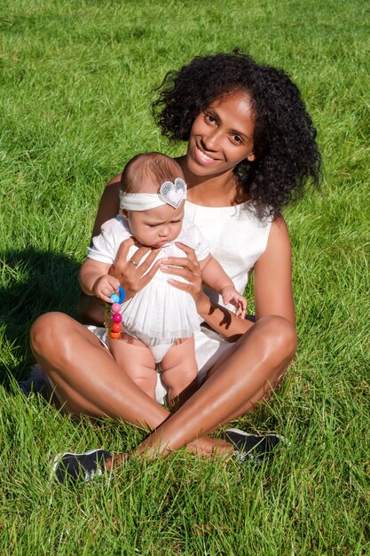 Young african american woman and her daughter sitting on green grass in summer park