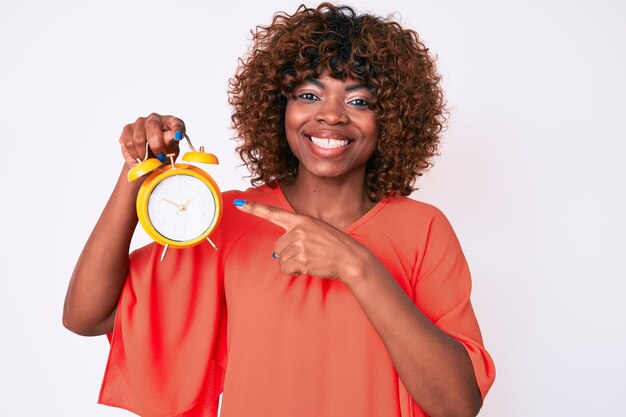 Young african american woman holding alarm clock smiling happy pointing with hand and finger