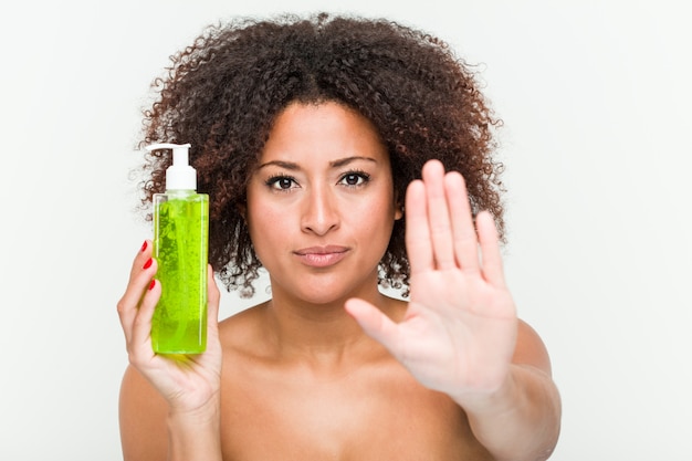 Young african american woman holding aloe vera bottle standing with outstretched hand showing stop sign, preventing you.