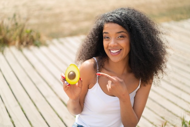 Photo young african american woman holding an avocado at outdoors and pointing it