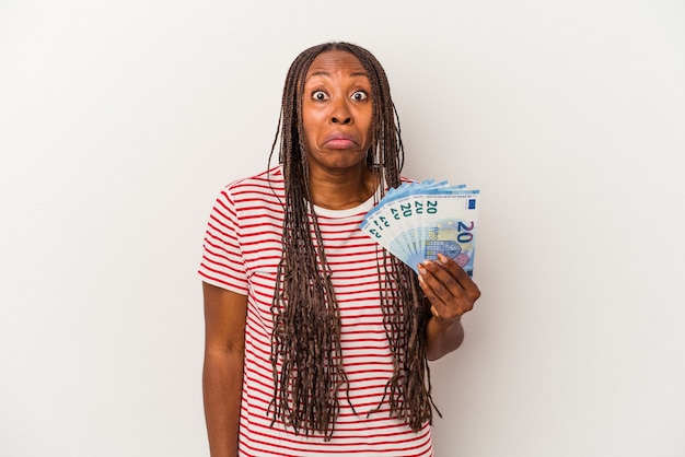 Young african american woman holding banknotes isolated on white background shrugs shoulders and open eyes confused.