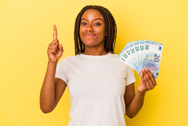 Young african American woman holding bills isolated on yellow background  showing number one with finger.