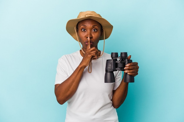 Young african american woman holding binoculars isolated on blue background keeping a secret or asking for silence.