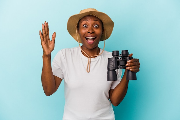 Young african american woman holding binoculars isolated on blue background receiving a pleasant surprise, excited and raising hands.