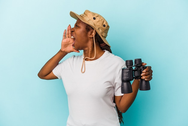 Young african american woman holding binoculars isolated on blue background shouting and holding palm near opened mouth.