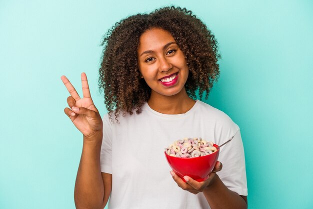 Young african american woman holding a bowl of cereals isolated on blue background joyful and carefree showing a peace symbol with fingers.