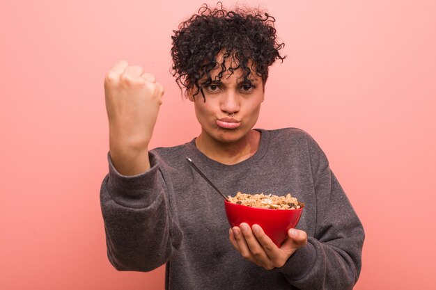 Giovane donna afroamericana che tiene una ciotola di cereali che mostra pugno, espressione facciale aggressiva.