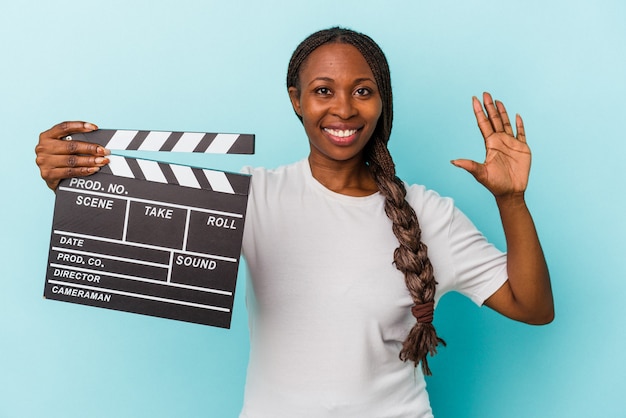 Young african american woman holding clapperboard isolated on blue background smiling cheerful showing number five with fingers.