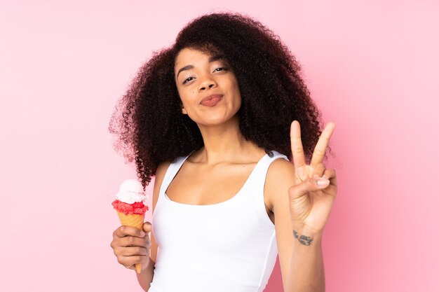 Молодая афро-американская женщина, держащая мороженое корнет, улыбается и показывает знак победы