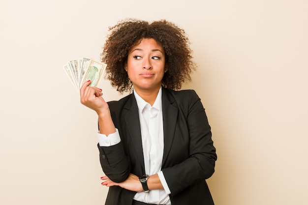 Young african american woman holding dollars looking sideways with doubtful and skeptical expression.